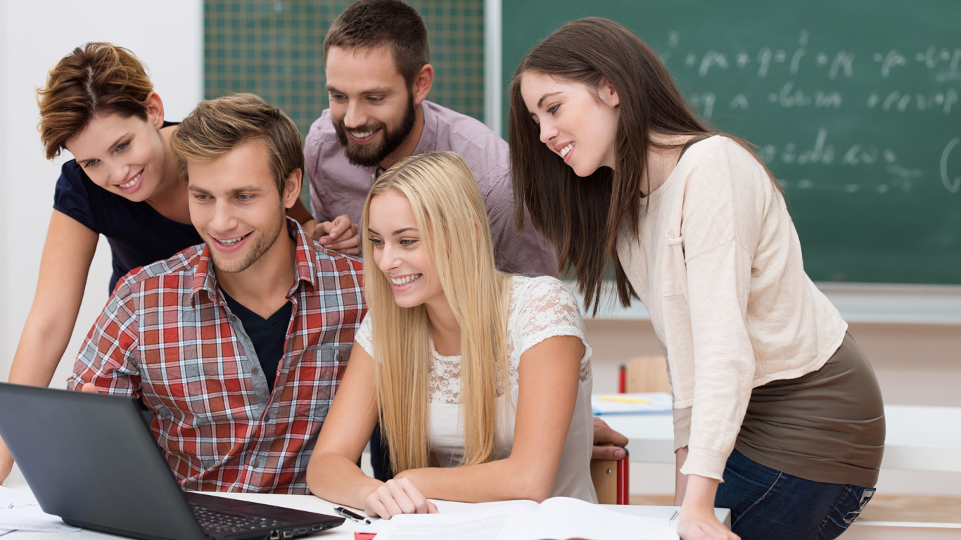 Successful team of diverse young male and female students in the classroom smiling with delight as they read the outcome of their project on a laptop computer